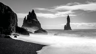 Black and white photo of a misty ocean shore with sharp, jagged rock formations rising from the water under a cloudy sky