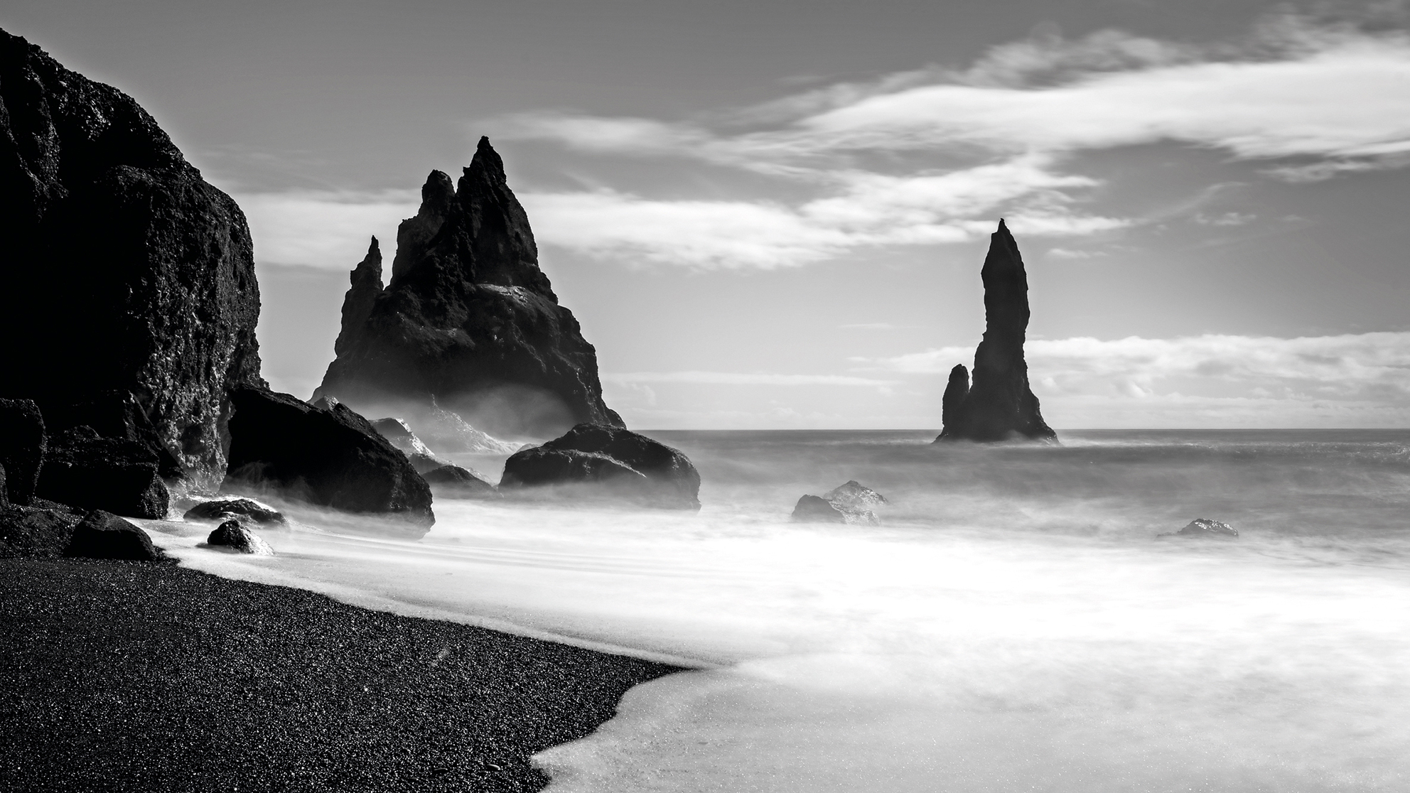 Black and white photo of a misty ocean shore with sharp, jagged rock formations rising from the water under a cloudy sky
