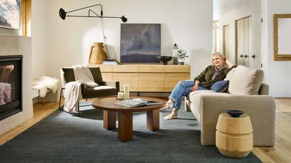 modern living room with navy blue rug, Emily Henderson sits on the couch, round timber coffee table, teak console with art and fireplace