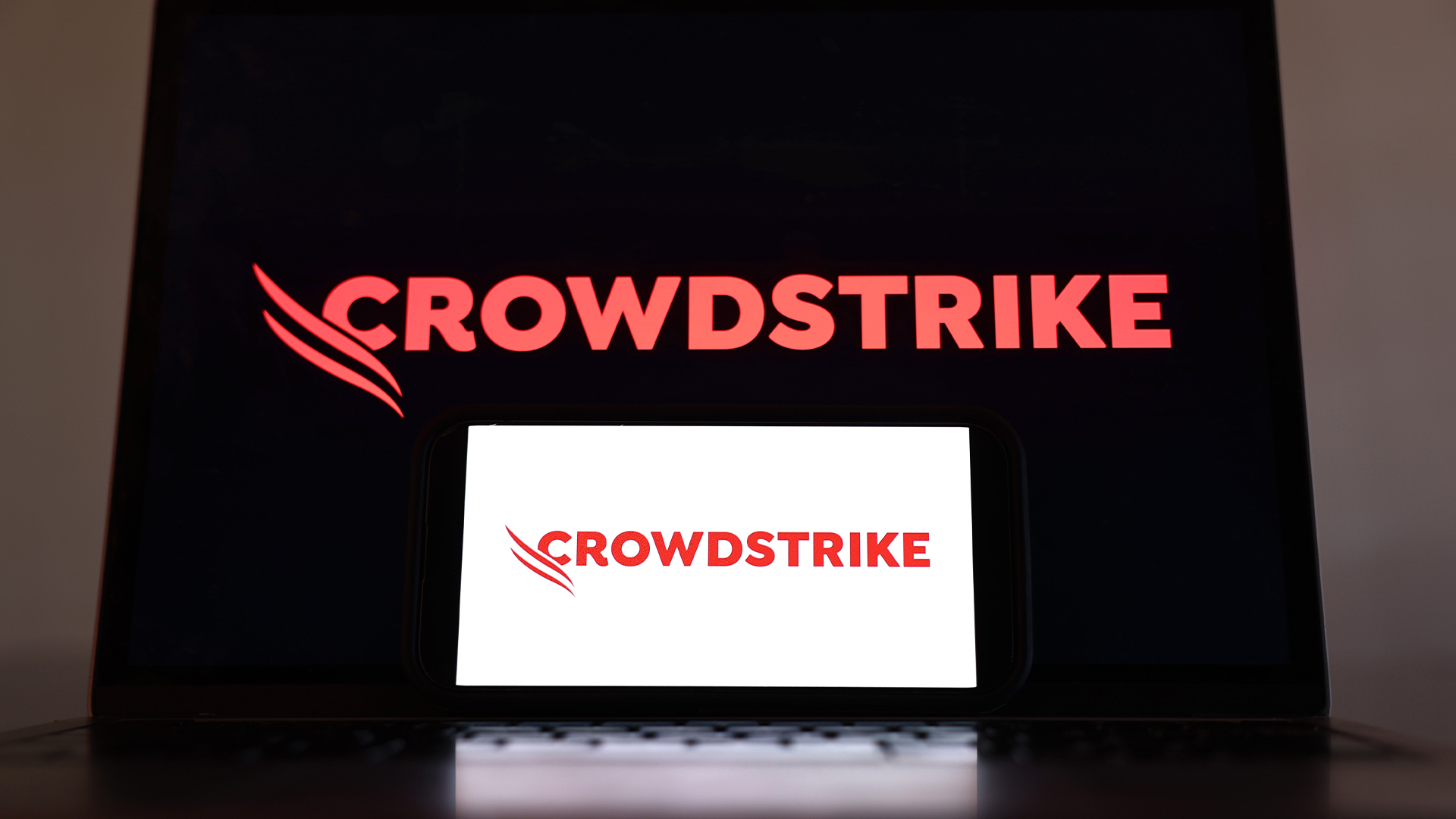 CrowdStrike logo and branding pictured on a tablet screen in red lettering on a black background, with logo on smartphone screen in foreground in red lettering with white background.