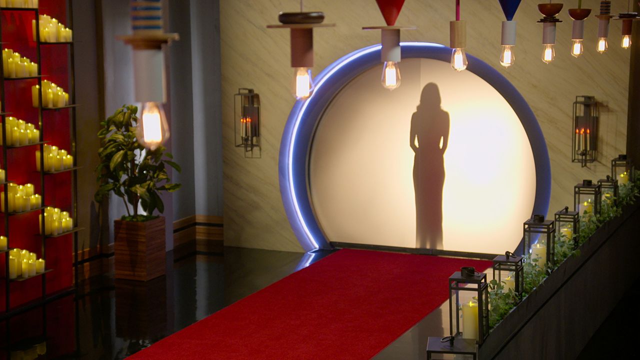 A woman stands behind a backlit screen, seen from a room decorated with a red carpet, handing Edison bulbs, and candle displays, in &#039;Love is Blind&#039; season 7.