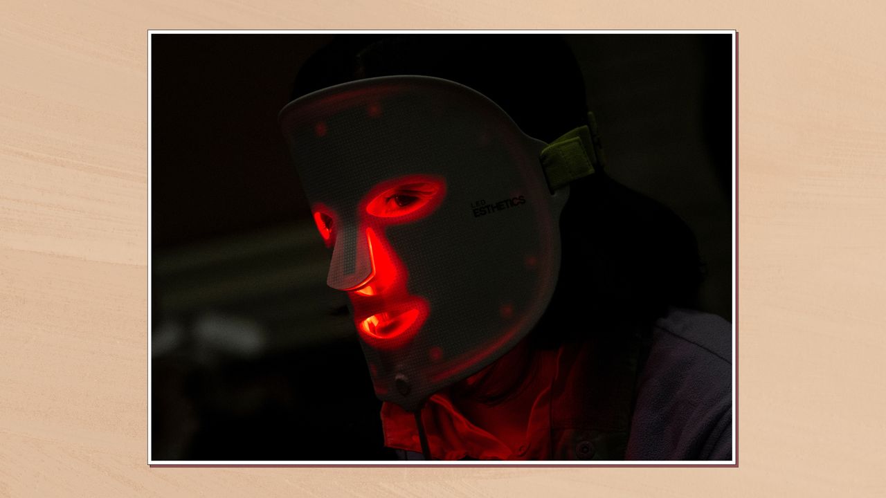 Image of woman wearing a red light therapy mask while in the dark, in a white frame against a beige watercolour-style background 