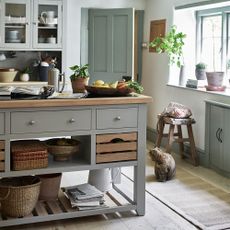 a grey freestanding kitchen island with draws and shelves and a wooden top, in the middle of a kitchen with grey cabinets and green door