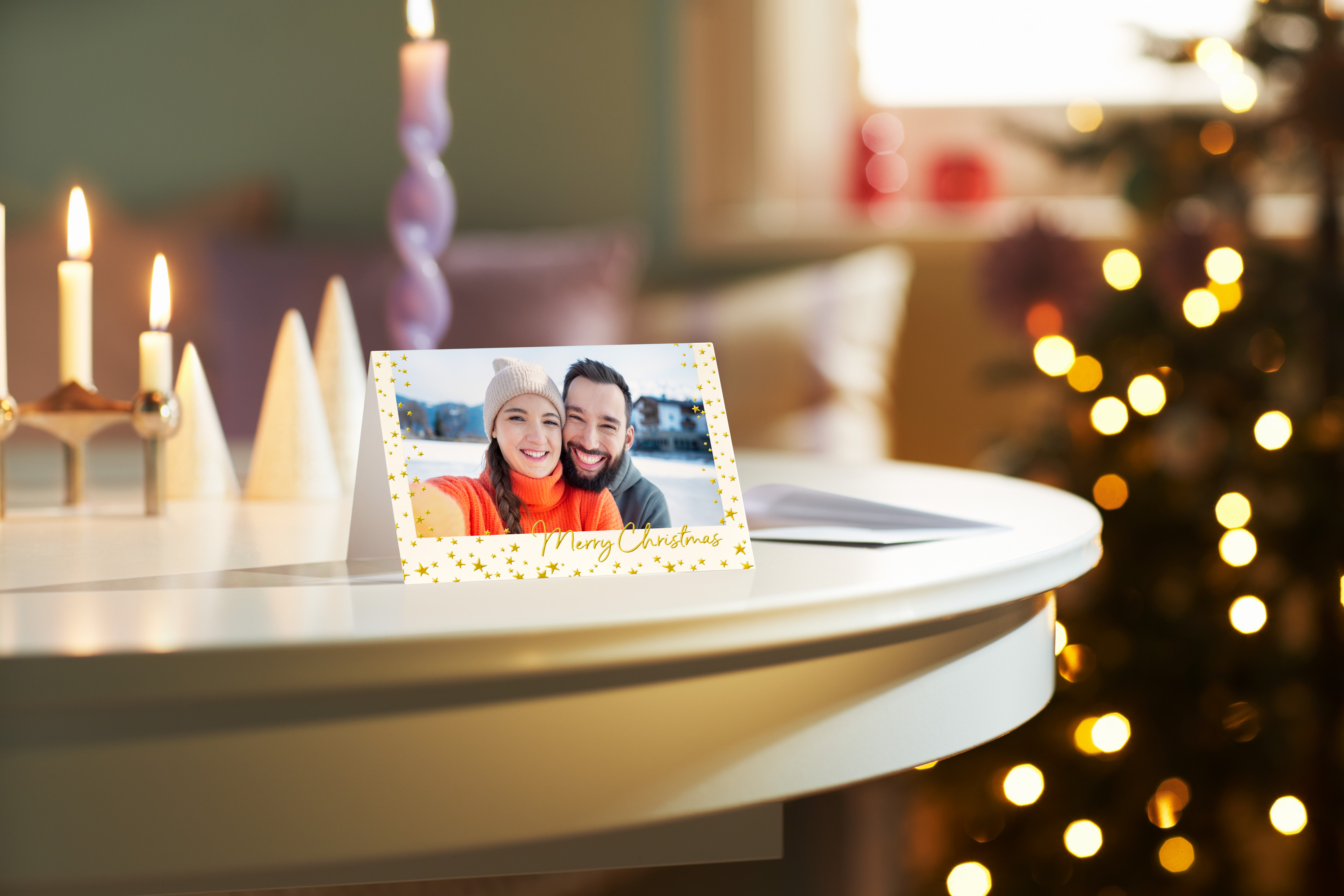 A printed Christmas card featuring a photo of a smiling couple in winter hats is displayed on a white table next to lit candles, with a blurred Christmas tree in the background.