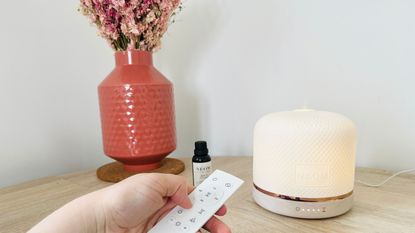 The Neom Wellbeing Pod Luxe essential oil diffuser on a side with some dried flowers and essential oils, holding the remote 