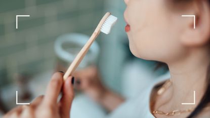 Woman using white toothbrush in bathroom mirror to whiten teeth naturally