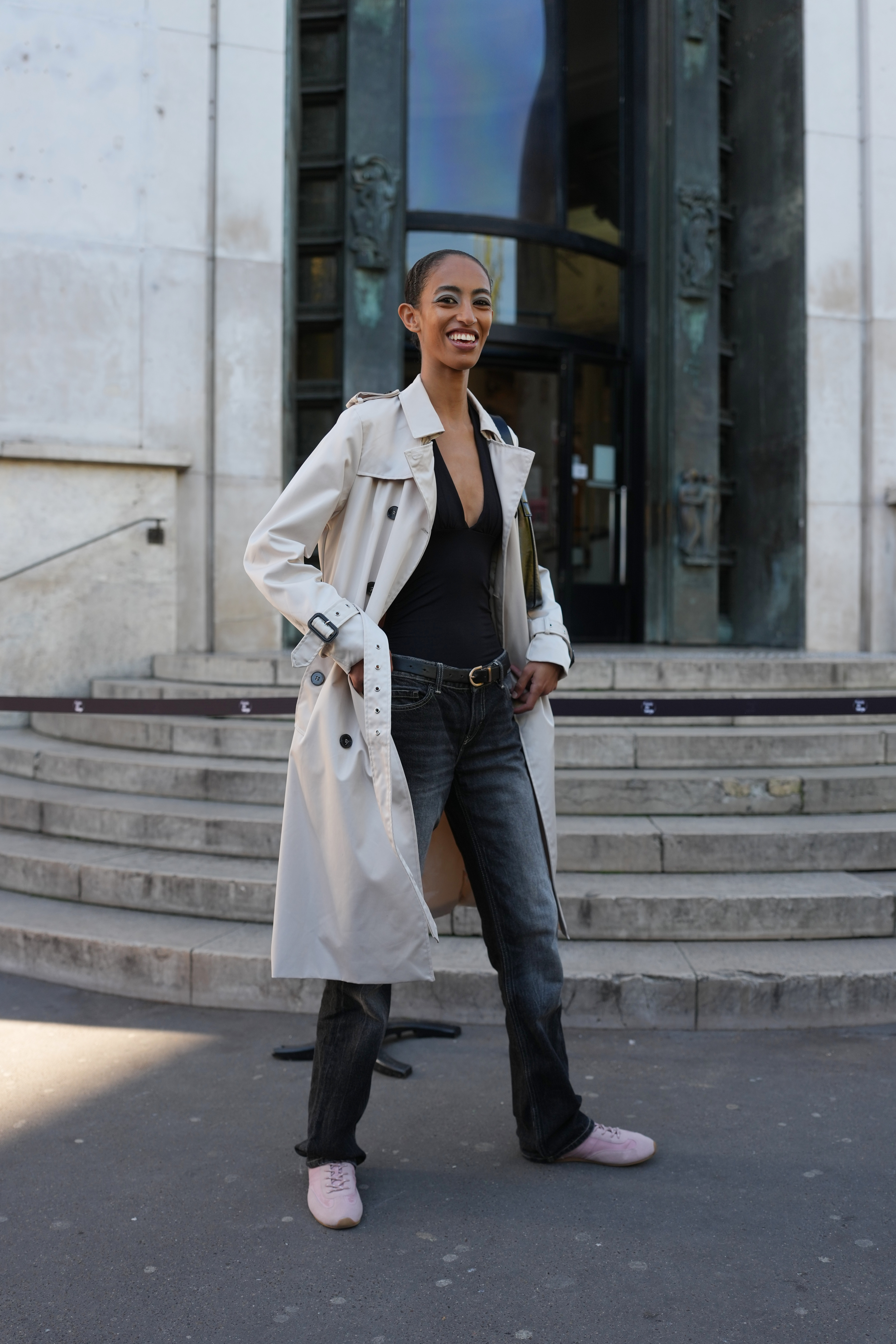 PARIS, FRANCE - OCTOBER 07: A model wears hair pulled back into a low slick bun, dark brown hair, a black leather shoulder bag, a light beige trench coat with double-breasted buttons, epaulettes and belted cuffs worn over a black deep V-neck top, faded black denim jeans pants with a black leather belt with a gold metal buckle, and pink sneakers shoes, outside Kiko Kostadinov, during Paris Fashion Week - Womenswear Spring Summer 2026, on October 07, 2025 in Paris, France (Photo by Edward Berthelot/Getty Images)