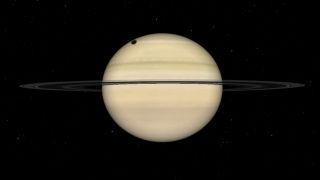 A simulation of Saturn is shown, with the dark silhouette of the large moon Titan visible as a dark circle on the upper left part of the planet's disk. Cloud bands are pictured crossing the planet's surface above and below the gas giant's ring system, which is orientated almost edge-on to the viewer.
