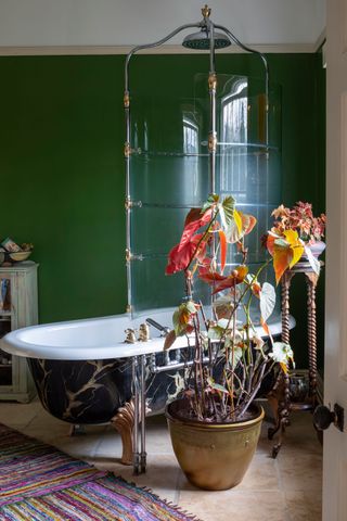 bathroom with green walls and roll top bath with marble effect sides and period style shower enclosure and large plant and rag rugs on limestone style floor tiles