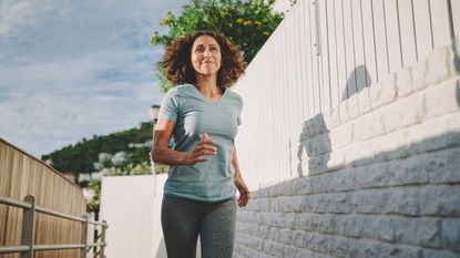 A woman in a t-shirt and leggings runs down a brightly lit alleyway. She is smiling and it is a sunny day.