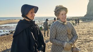 Synnøve Karlsen in a black bonnet and stands on a beach with Phyllis Logan in a beige bonnet and dress as Mrs Austen in Miss Austen.