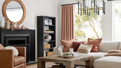 Terracotta cushions on a white sofa in front of windows looking onto a garden view; a mirror above a black fireplace with black bookshelves and a tan leather armchair either side. 