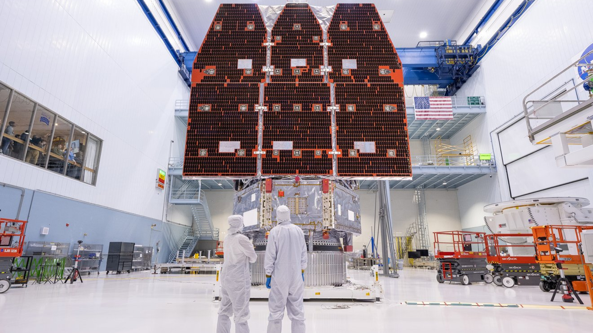 Three large solar panels hang in the back of a cleanroom warehouse room where two workers dressed in white suits stand in the foreground