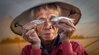 Elderly person in a conical hat playfully holds small fish over their eyes, mimicking glasses, with a joyful expression against a textured background