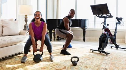 woman and man dressed in workout gear picking up a kettlebell each and smiling. they're in a living room with an exercise bike next to them, a sofa on the side and a piano in the background.