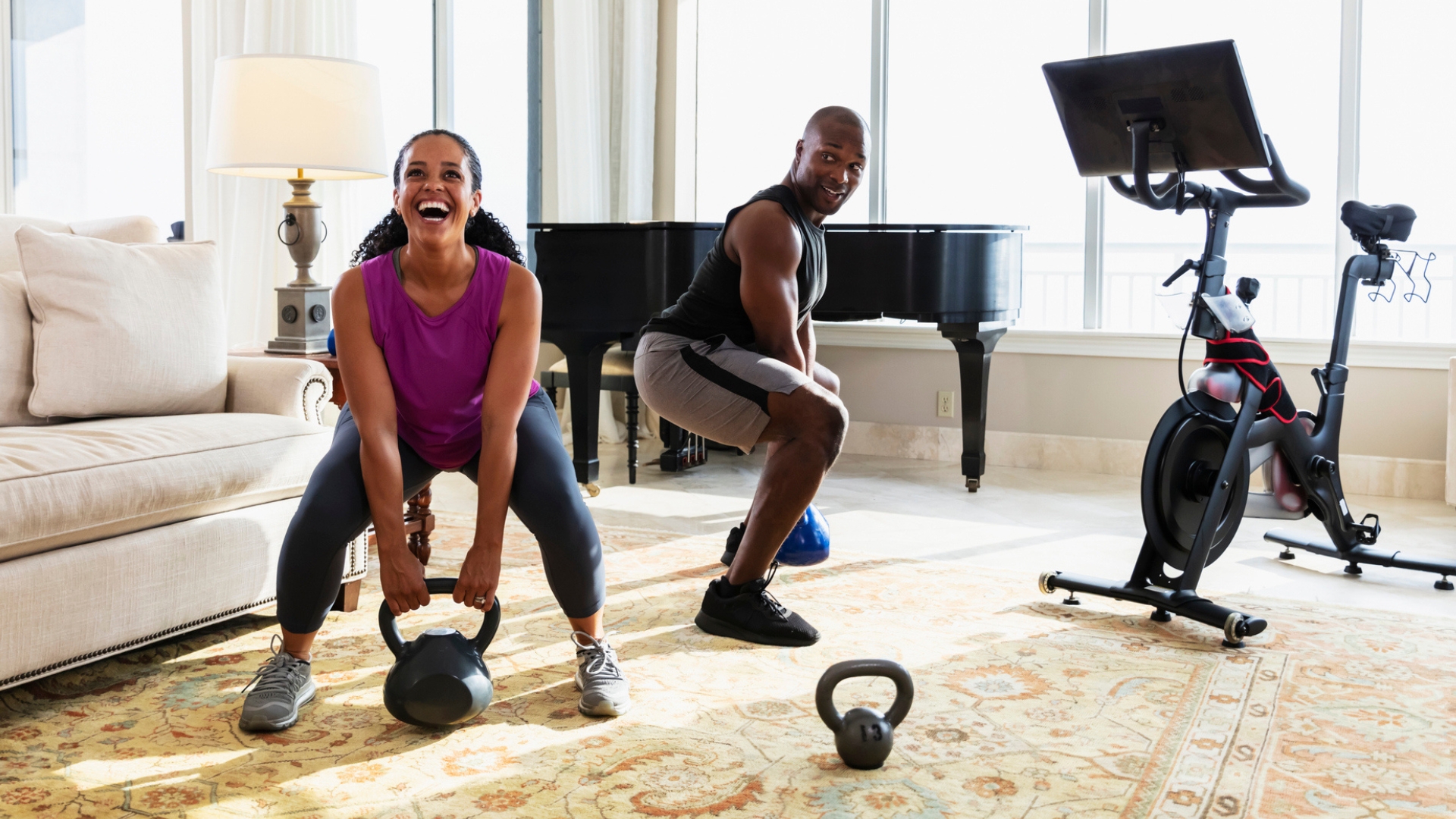 woman and man dressed in workout gear picking up a kettlebell each and smiling. they're in a living room with an exercise bike next to them, a sofa on the side and a piano in the background. 