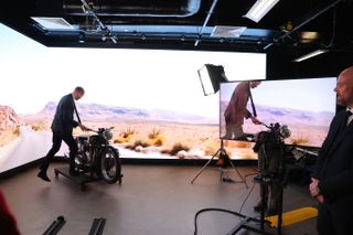 Prince William climbing on a motorcycle in front of a green screen with a road and mountain backdrop as lights shine on him and a TV