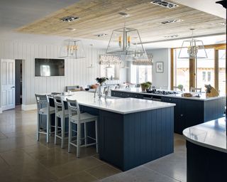 Large kitchen with two blue kitchen islands, white marble countertops, gray paneled walls, light wooden paneled ceiling section above the islands, four glass pendants, dark gray stone flooring
