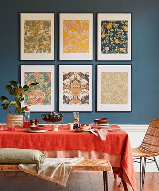 Dining table with red tablecloth, bench seat and chair on wooden floor, dark blue wall in the background with set if floral patterned pictures, framed wallpaper panels.