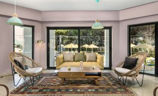 Interior view of a seating area at The Magnolia Hotel featuring light coloured walls, pastel green pendant lights, a multicoloured patterned rug, two wicker style chairs with cushions, a pale yellow sofa with cushions and glass doors with black frames leading to the outside area