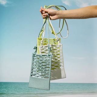 A hand is seen holding three different knitted bags against a sun-lit, seaside horizon