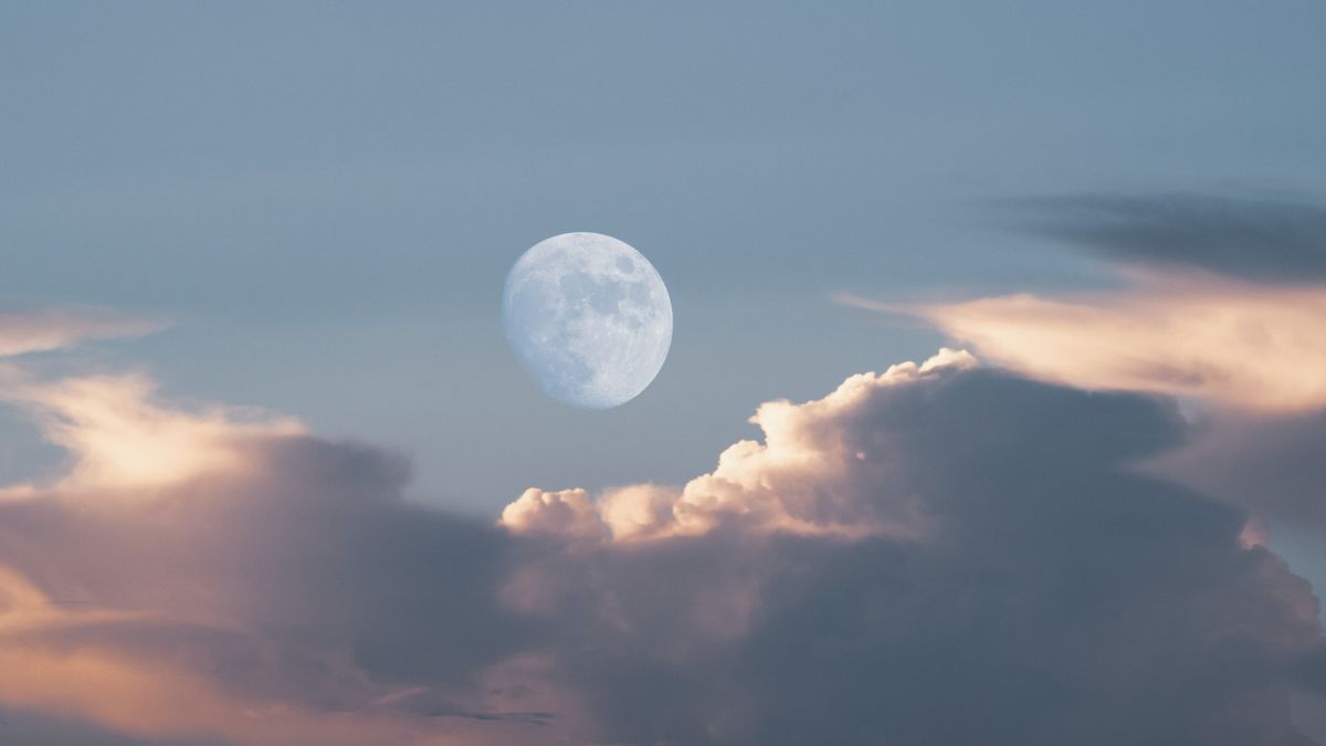 The nearly-full daytime moon hangs dimly in a blue sky near clouds