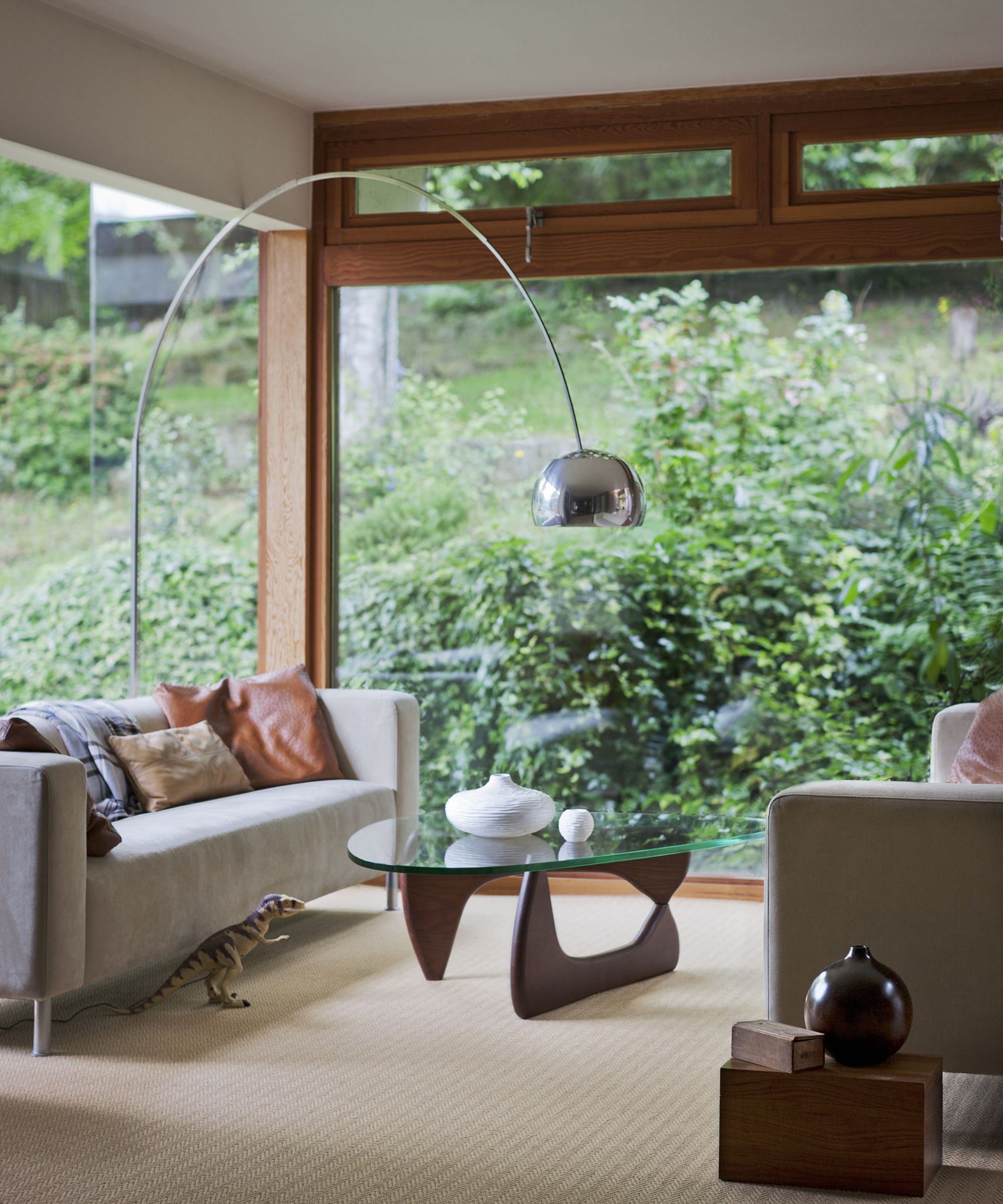 Arc floor lamp curves over glass and walnut coffee table between grey sofas facing a garden window.