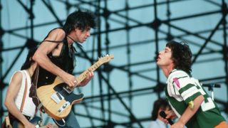 Mick Jagger, wearing a football jersey, interacts with Keith Richards during a Rolling Stones concert. Ron Wood plays guitar beyond