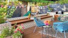Outdoor decor trends are so chic. Here is a green outdoor awning over a patio scene with black and white tiles, dark brown decking, potted plants, and a blue wicker set with three round seats and a coffee table with a black side table with colorful flowers