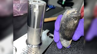 a gloved hand holds a burned piece of metal about the size and shape of a banana