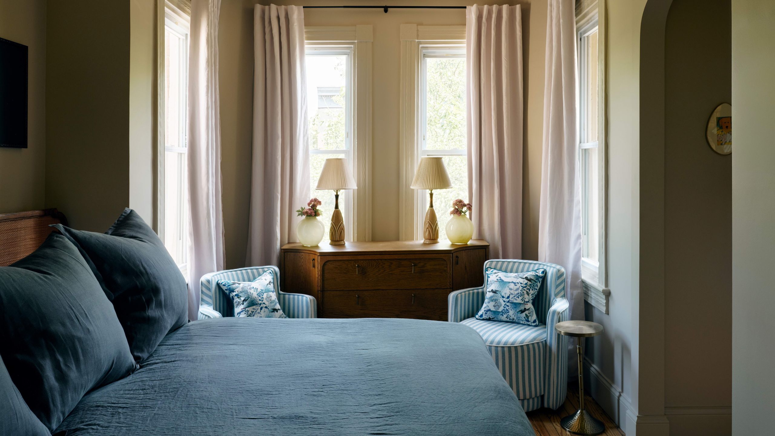 a large soft green primary with a bay window styled with drapes, wooden dresser, two blue striped chairs and a bed made with dusty blue linens