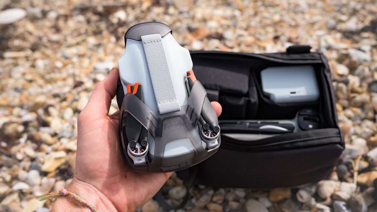 Potensic Atom 3 beginner drone in user's hands with its propellors folded away and the Fly More Combo bag and its content behind it, on a pebble beach