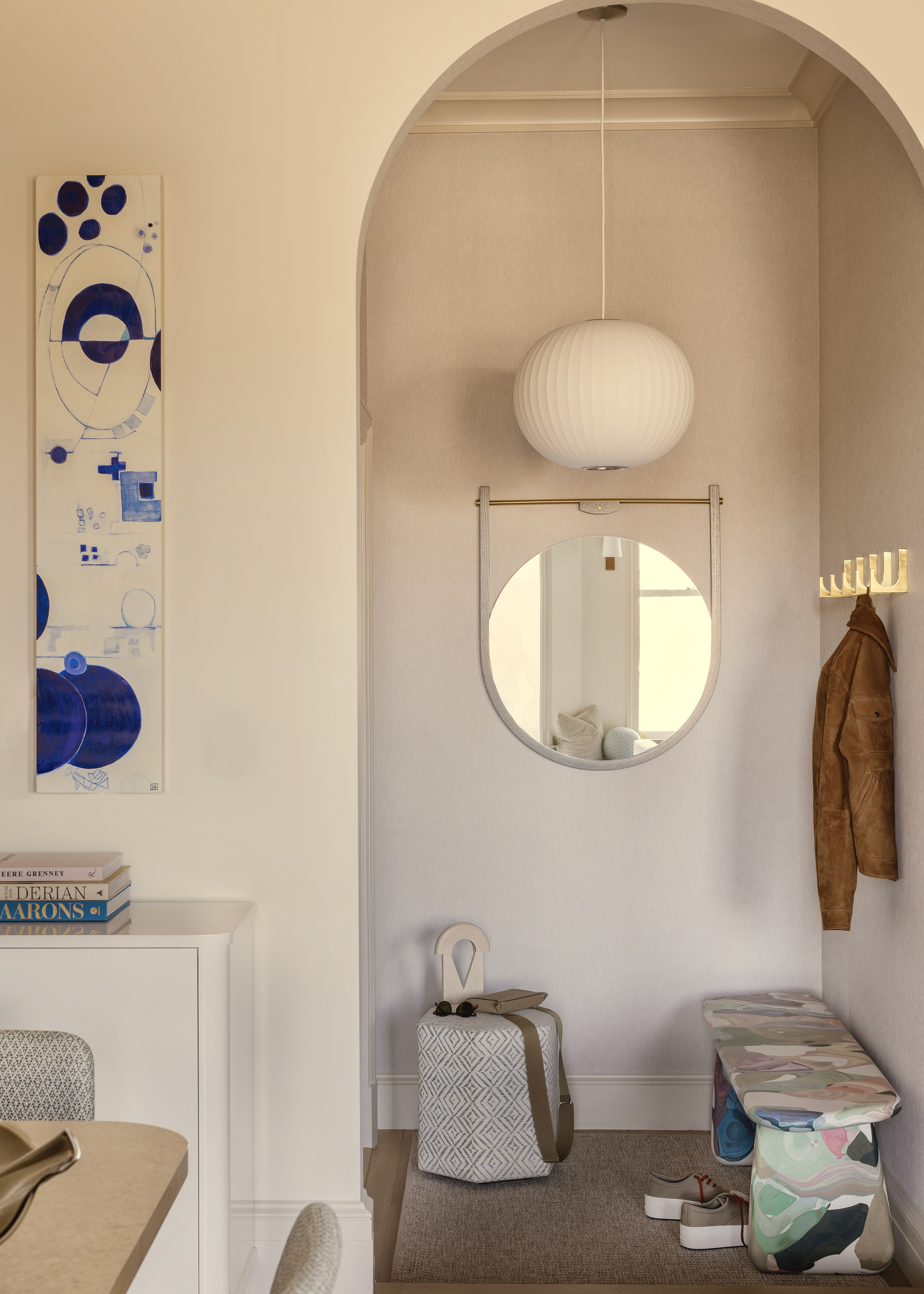 A small mudroom through an arched doorway with a unique and colorful bench made from recycled materials, a white pendant light above, coat hooks on the wall and a mirror shining light from the window opposite