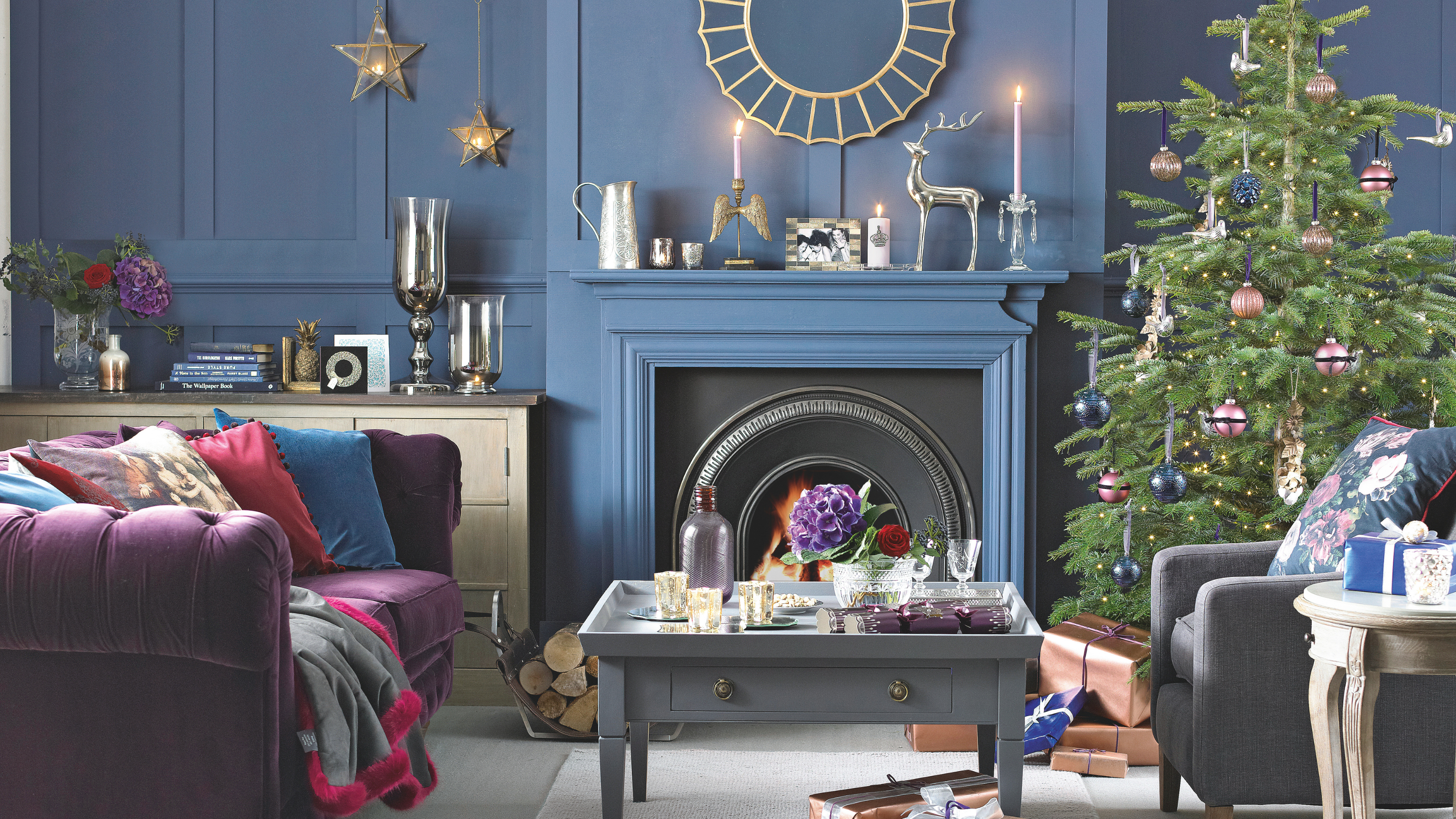 Blue living room with Christmas decoration, purple suede sofa, grey armchair, grey coffa table, decorated tree and fireplace.