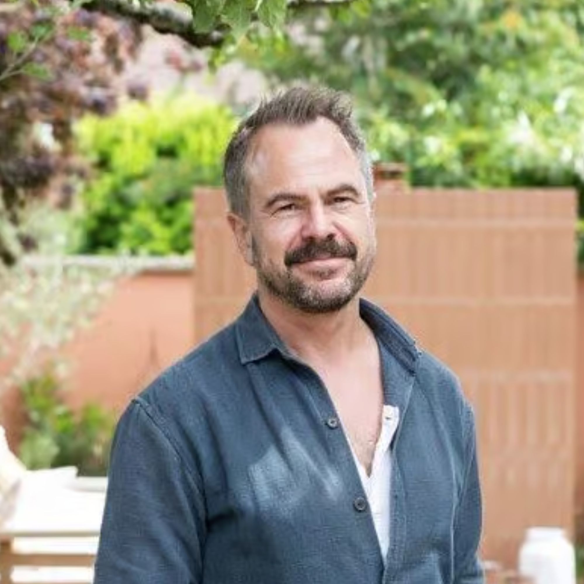 Man with grey beard in navy denim shirt smiling in a garden, blurred trees and wooden fence behind him.