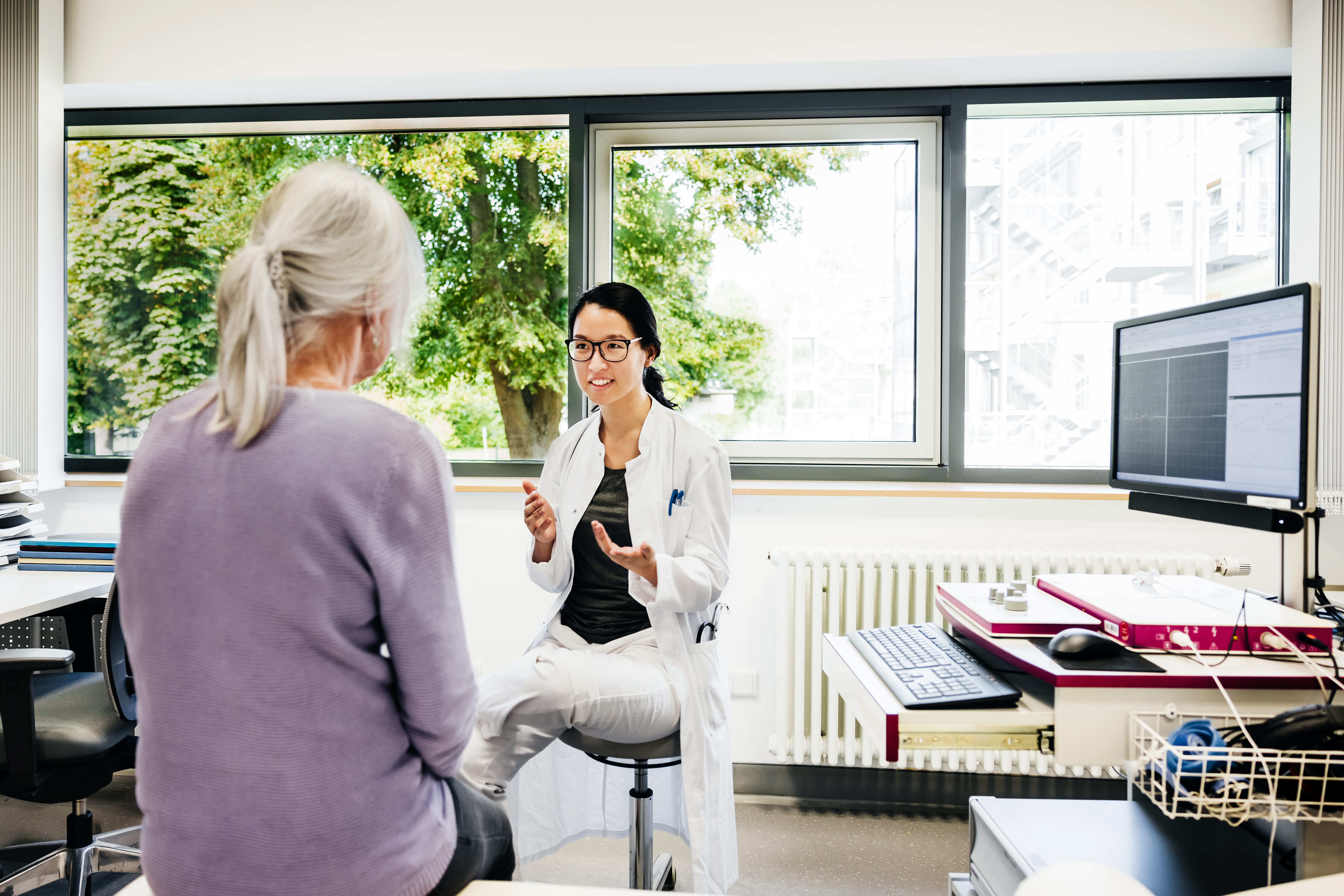 A young female doctor in a white coat and eyeglasses is seen gesturing during a conversation with an elderly patient in a doctor's office. The bright and airy room, with a large window showing a view of trees, suggests a friendly and open atmosphere.