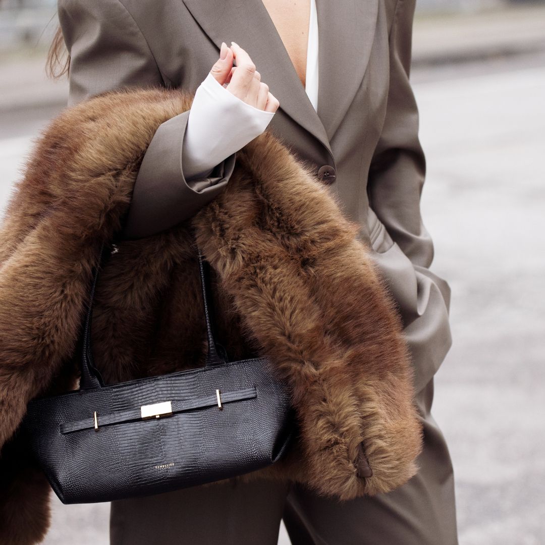 Fragrance trends Gili Biegun wears structured olive brown suit, holding a fur scarf, Demellier London The New York Shoulder east west bag in black lizard on December 06, 2025 in Copenhagen, Denmark