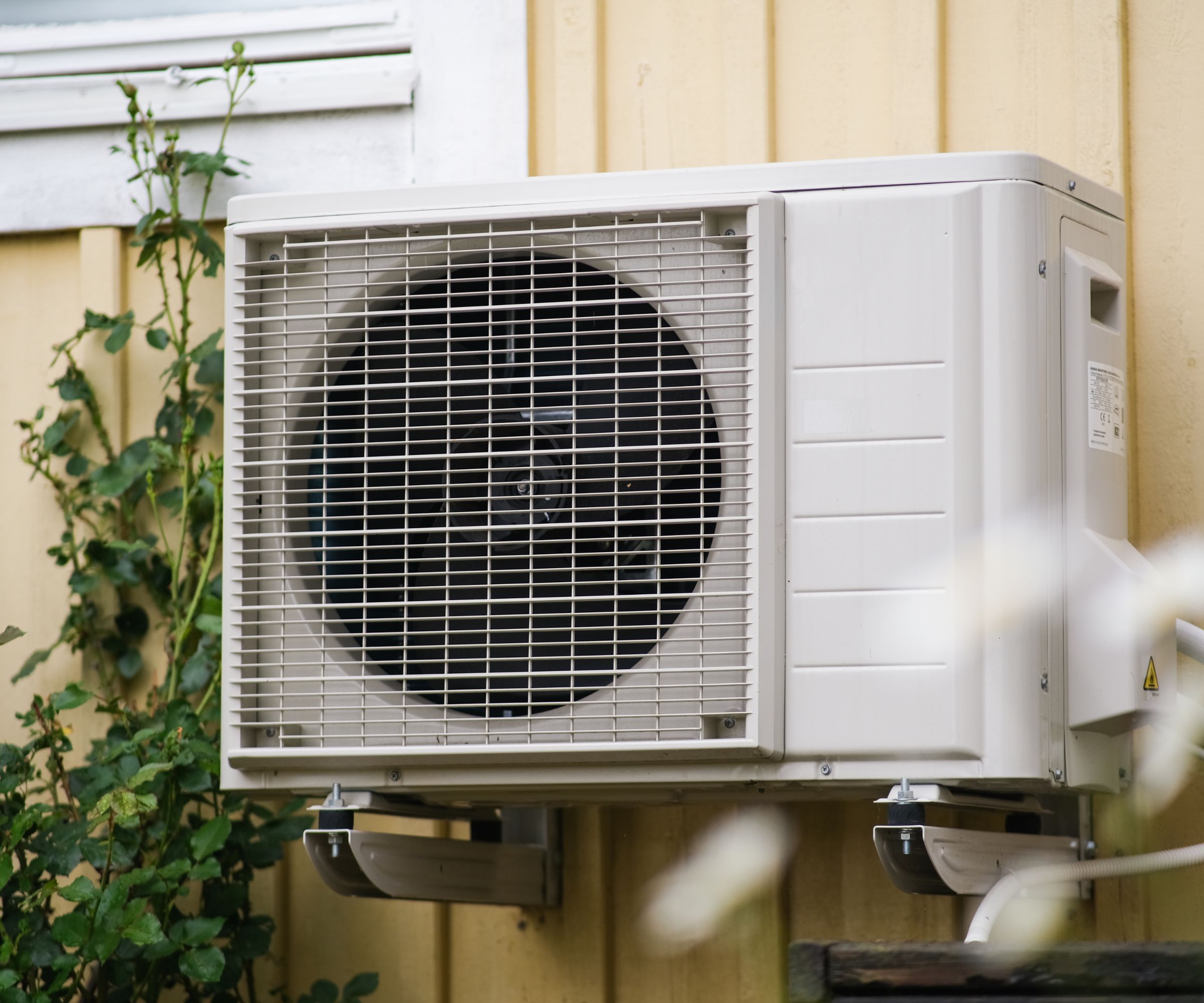 A white AC condenser unit mounted to the side of a yellow sided home.