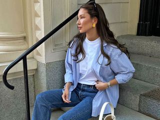 Fashion influencer Felicia Akerstrom poses on steps outside a building in Stockholm, Sweden wearing yellow gold earrings, a white t-shirt, blue striped button-down shirt, and jeans.