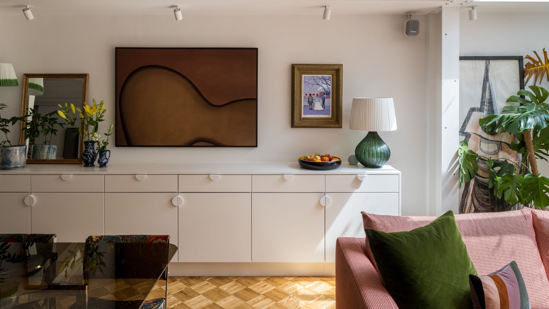 A living room with a white sideboard, checkerboard wooden flooring, a pink couch with green and striped cushions, and a dark amber table. Brown art and a framed painting are on the wall above the sideboard which holds a green lamp with a white shade, vases with flowers and a mirror