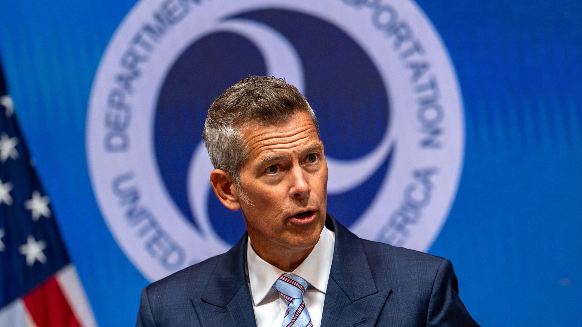 a man in a blue suit speaks in front of a plaque reading &quot;united states department of transportation&quot;