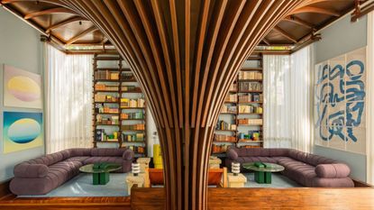 a living room with a purple sofa running its perimeter, a bookcase built into the wall, a timber funnel-shaped column in the center, blue walls, green coffee tables, and sheer curtains