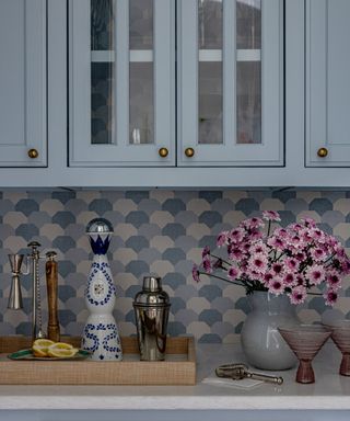 close up of light blue kitchen cabinets, white countertop with tray with decorative objects and cocktail shaker, vase of pink flowers on the side