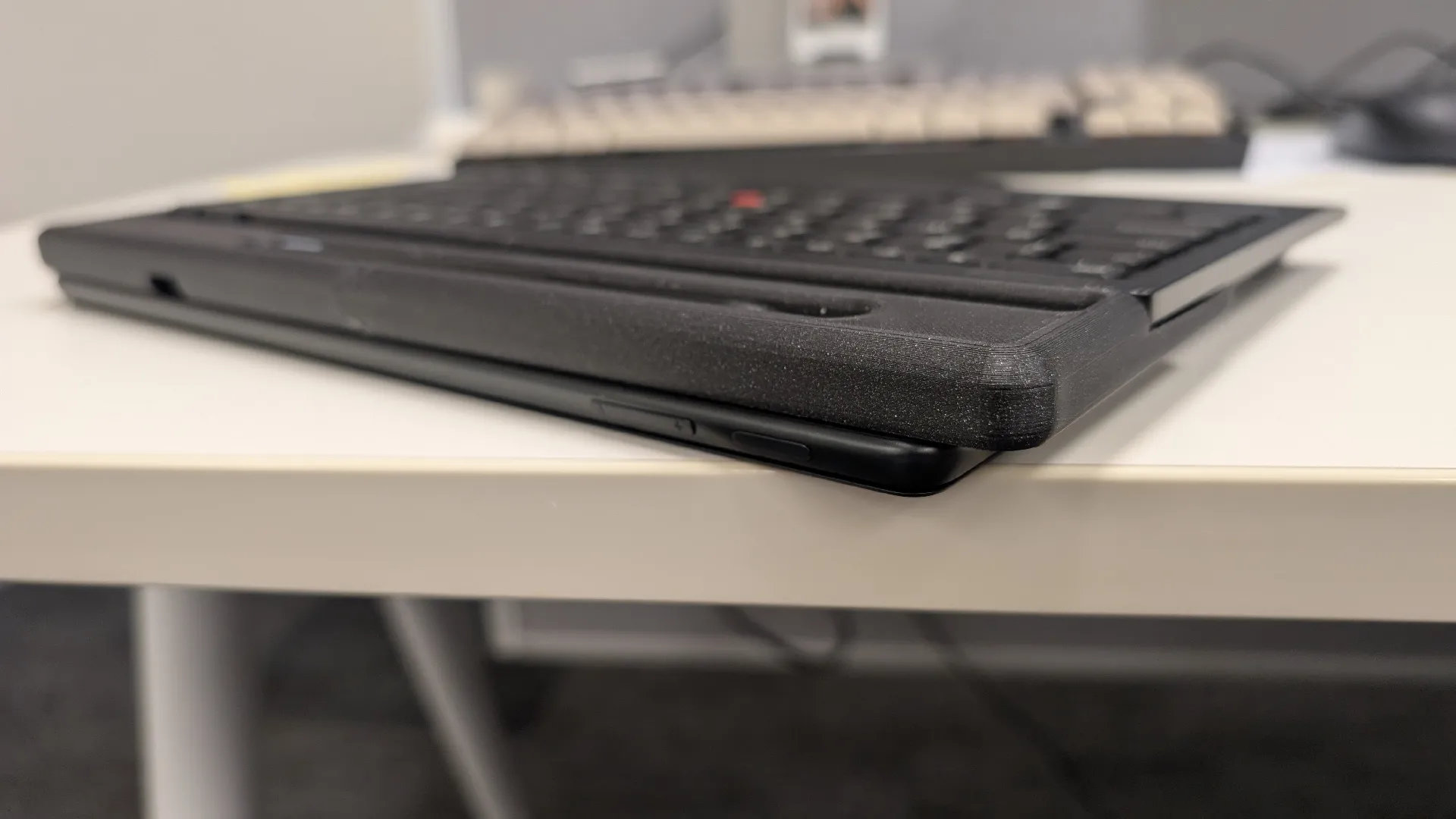 A closed black laptop with a keyboard cover on a white desk. In the background, out of focus, is another keyboard in an office setting, creating a professional atmosphere.