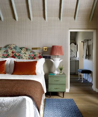 A fall color scheme in a bedroom with warm grey textured wallpaper, floral headboard, blue and white rug, green bedside table and burnt orange scatter cushions