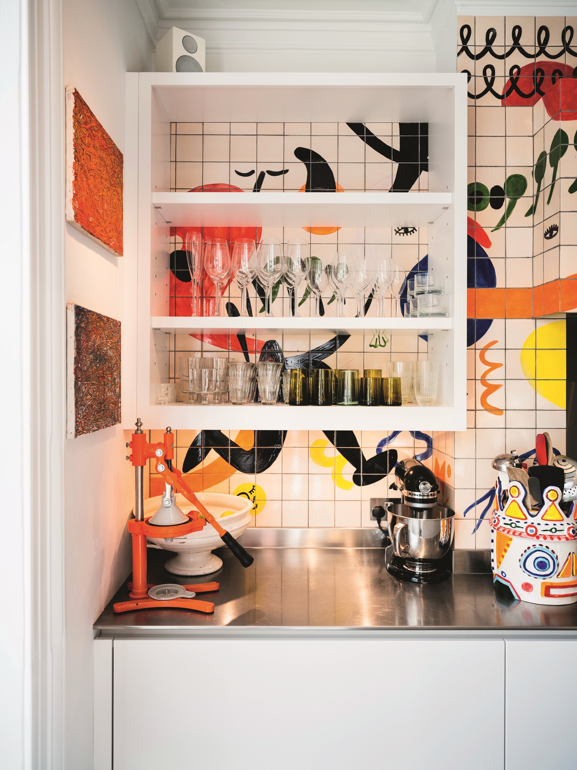 Yottam Ottolenghi's kitchen with floating white shelves, stainless counter, juice press, mural backsplash, and artwork on wall