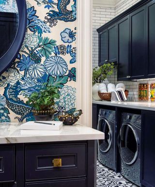 A navy blue mudroom laundry area with tiled flooring and walls, navy cabinetry, round mirror and printed blue wallpaper