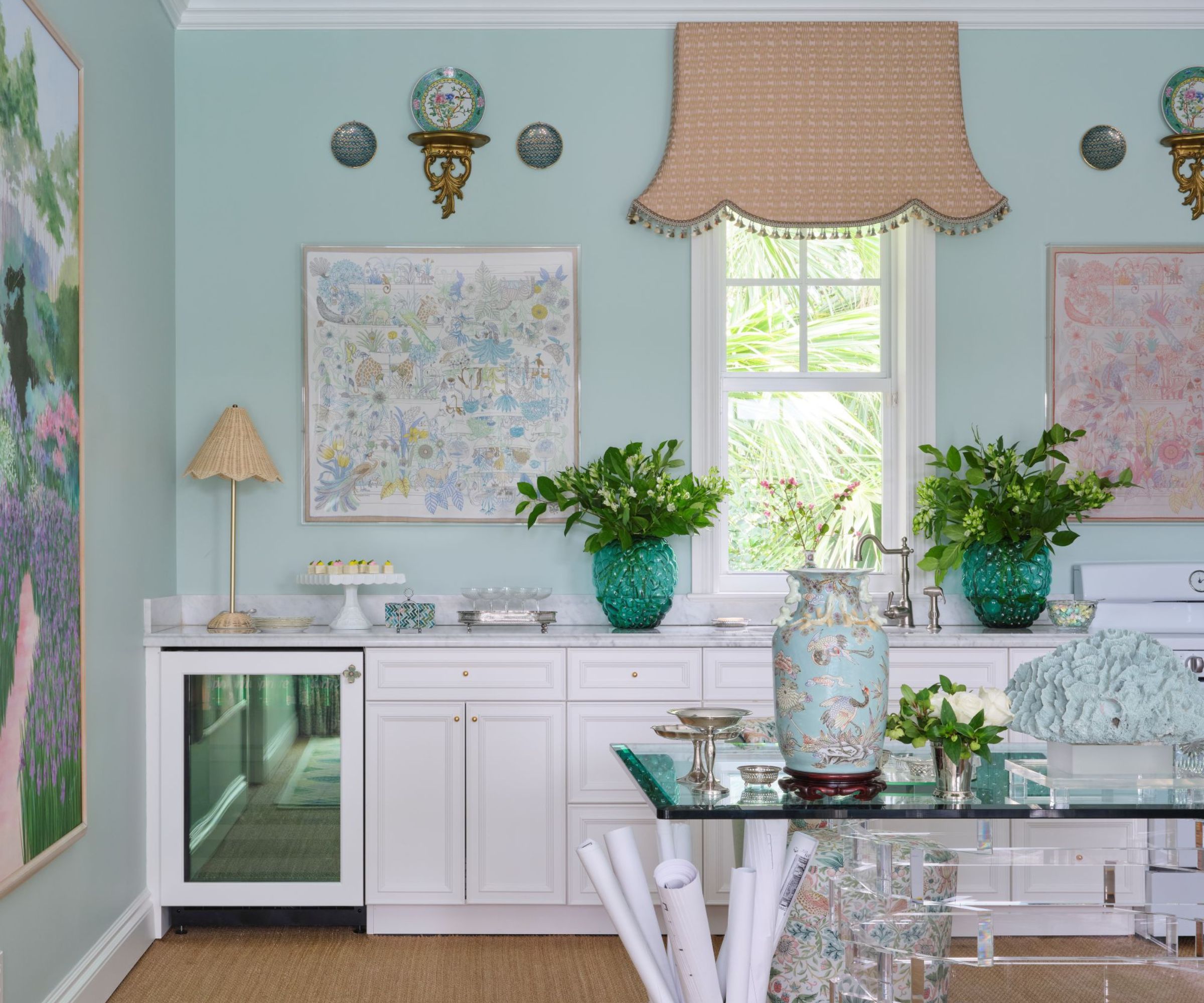 Home office with pale blue walls, rattan blind on window, glass desk and white cabinetry