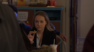 Amy Mitchell glares at Gina Knight and Dean Wicks in McKlunky's while eating a chicken nugget in EastEnders 