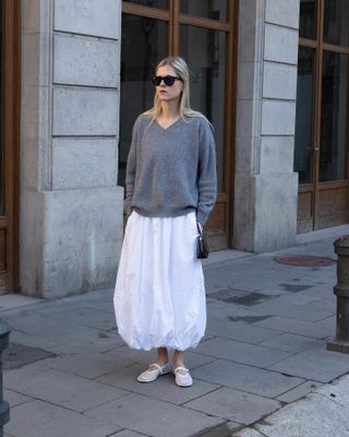 Influencer standing on street wearing gray V-neck sweater, white bubble skirt, and white ballet flats with black sunglasses.